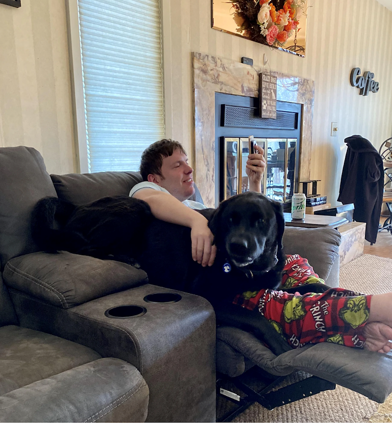 Un jeune homme en pantalon de pyjama rouge sourit devant son téléphone, allongé sur un canapé d'angle gris, un grand chien noir couché contre lui