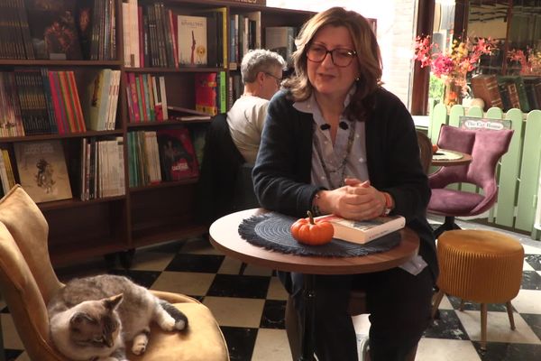 Sandra Valageas assise à une table de sa librairie, un chat gris et blanc couché sur un fauteuil moutarde au premier plan