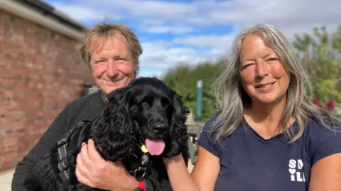 Rob Grubb et Jane Stuart Brown souriants, Hero contre eux devant un mur de briques