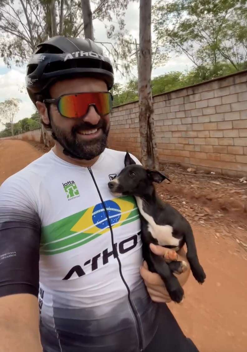 Le cycliste casqué sourit en tenant le chiot noir et blanc contre son torse, sur un chemin de terre rouge
