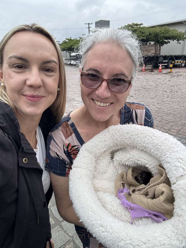 Deux femmes souriantes, l'une tenant un coussin blanc où repose le chaton emmailloté